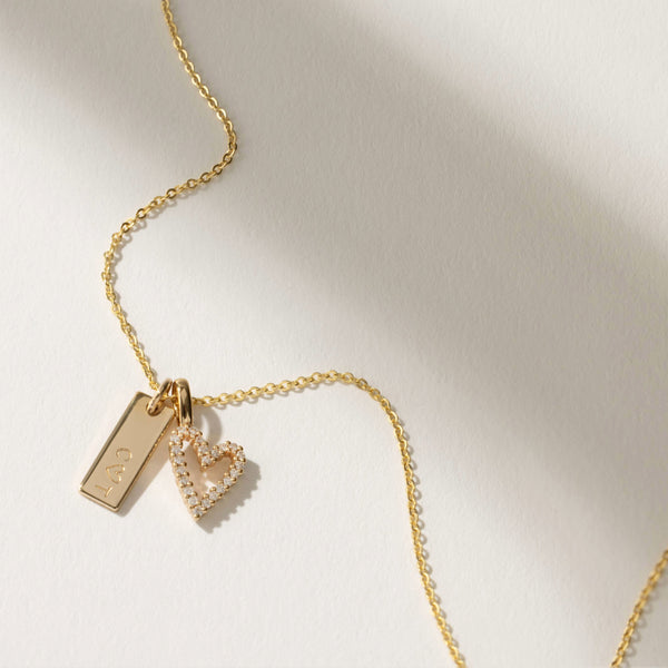A delicate gold pendant chain with an engraved mini rectangular bar charm and a heart outline charm covered in clear accent stones, set against a white background with shadows.