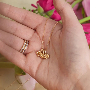 A person holding a delicate gold chain with multiple gold flower charms, shown in front of a bouquet of pink and white flowers.