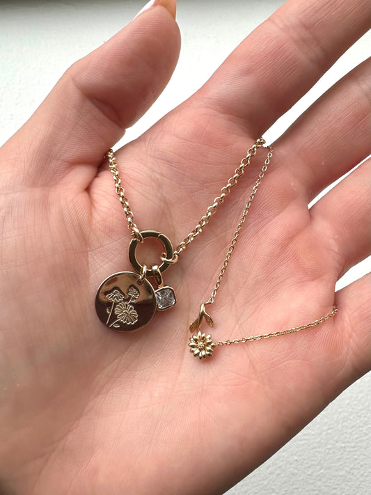 A hand holding a gold charm necklace with a clear gemstone and round flat gold disc charm, and a gold necklace with a horizontal daisy flower.