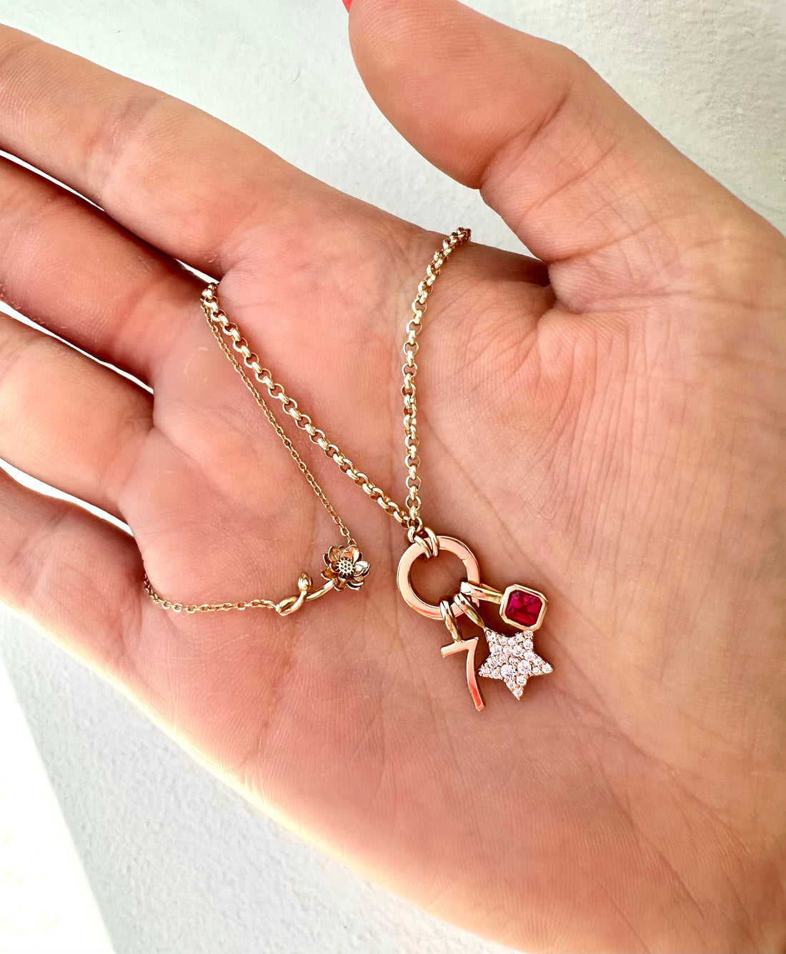 A hand holding a gold chain necklace with a lotus flower and a gold charm necklace with a red gemstone charm.