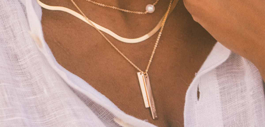 A woman wearing a thick gold chain necklace layered with a gold pendant chain featuring two different sized vertical bar charms.
