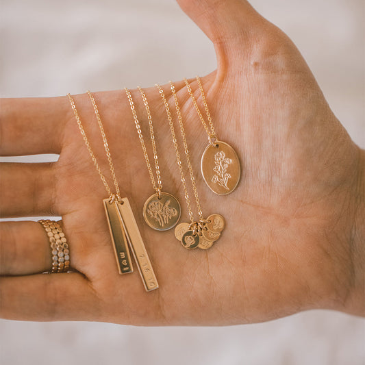 A hand holding four pendant chain necklaces, each with a different style of stamped charm.