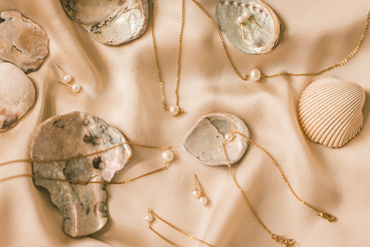 Multiple pieces of gold jewelry with round pearls laying on a cream satin surface with seashells.
