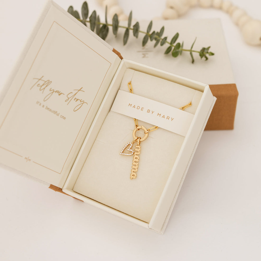 An open Made by Mary branded jewelry box with a gold charm necklace, featuring a heart and 'mama' charm.
