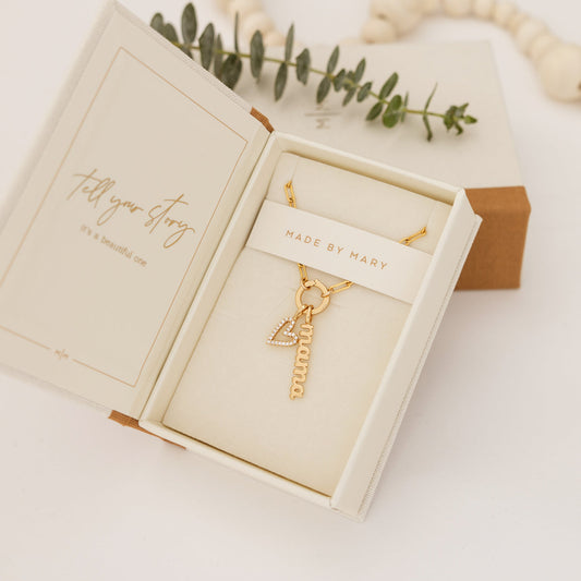 An open Made by Mary branded jewelry box with a gold charm necklace, featuring a heart and 'mama' charm.