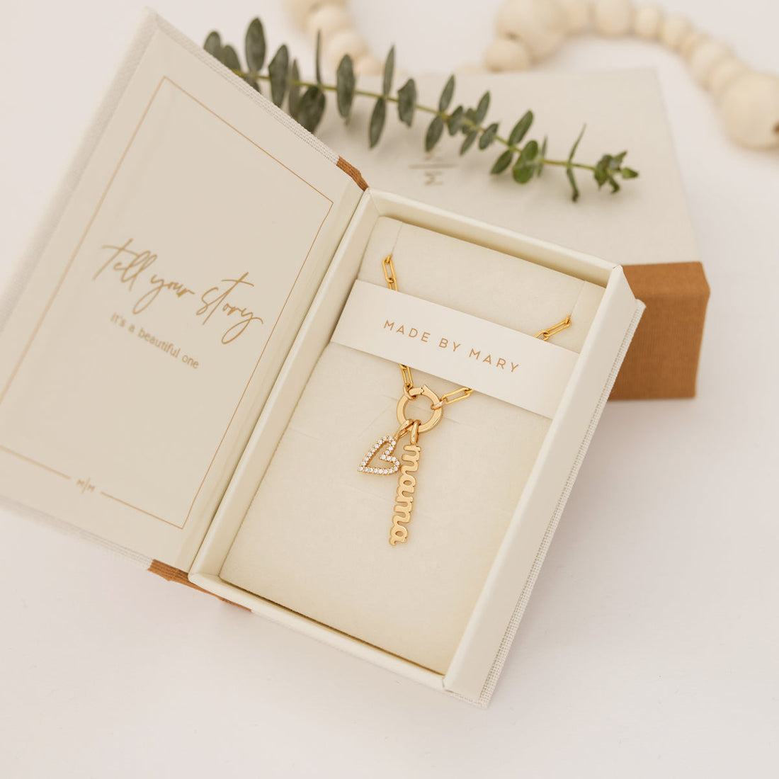 An open Made by Mary branded jewelry box with a gold charm necklace, featuring a heart and 'mama' charm.