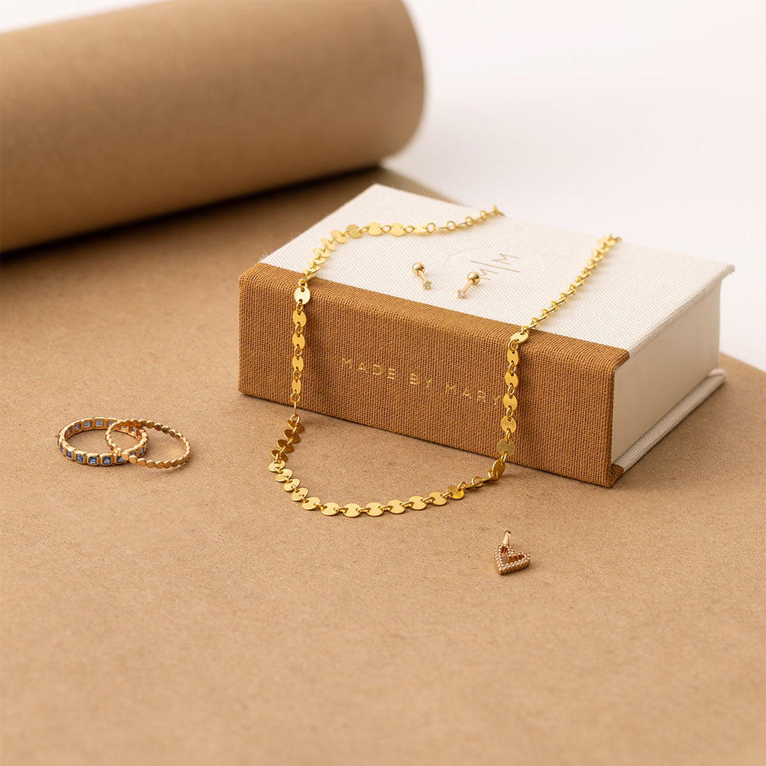 A Made by Mary book box with a gold necklace and gold studs on it, sitting beside two gold rings.