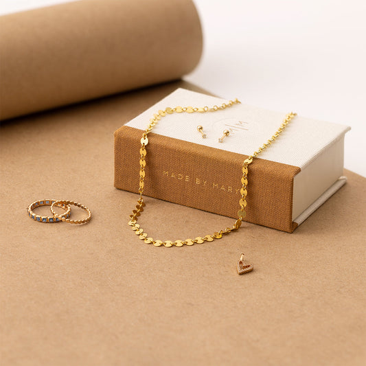 A Made by Mary book box with a gold necklace and gold studs on it, sitting beside two gold rings.