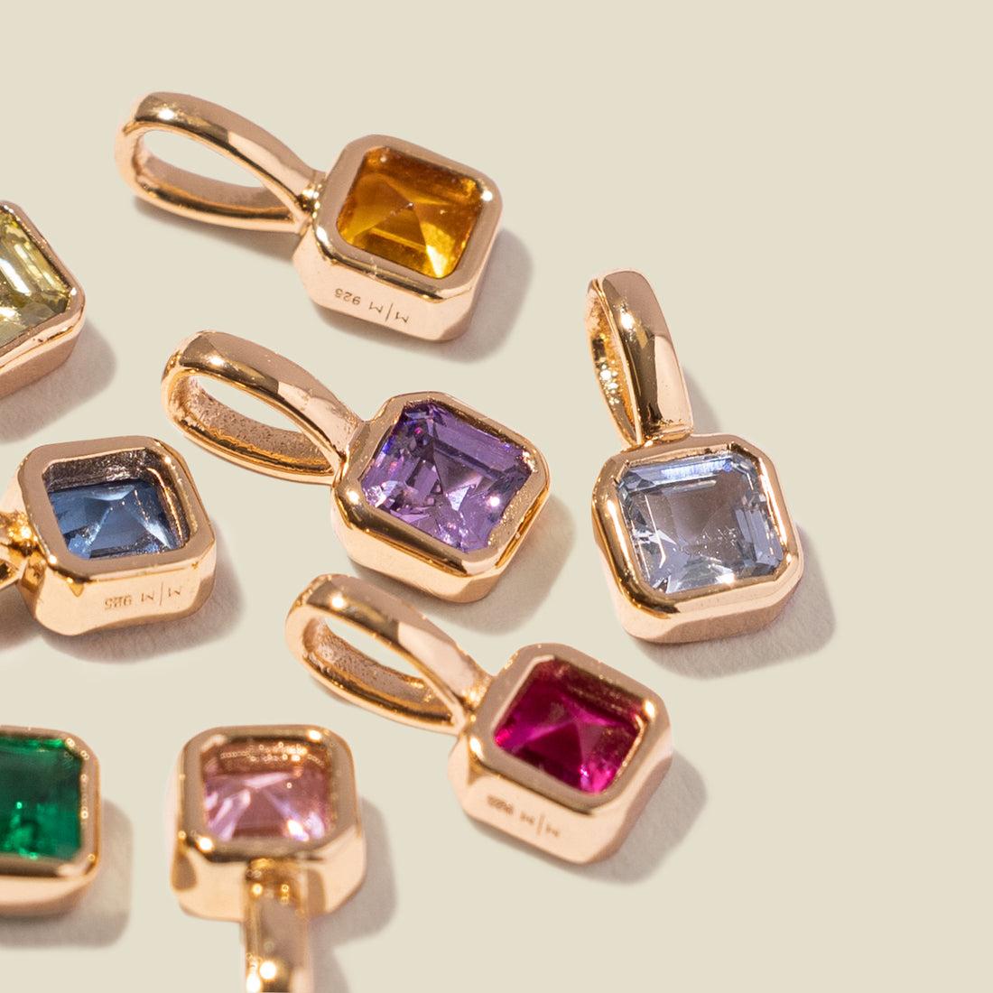 Multiple gold charms laying on a solid light beige surface, each with a different color of square gemstone.