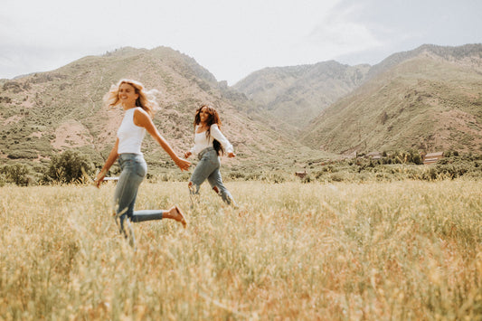 Two women frolicking in a field of beige grass in a meadow in front of a mountain.