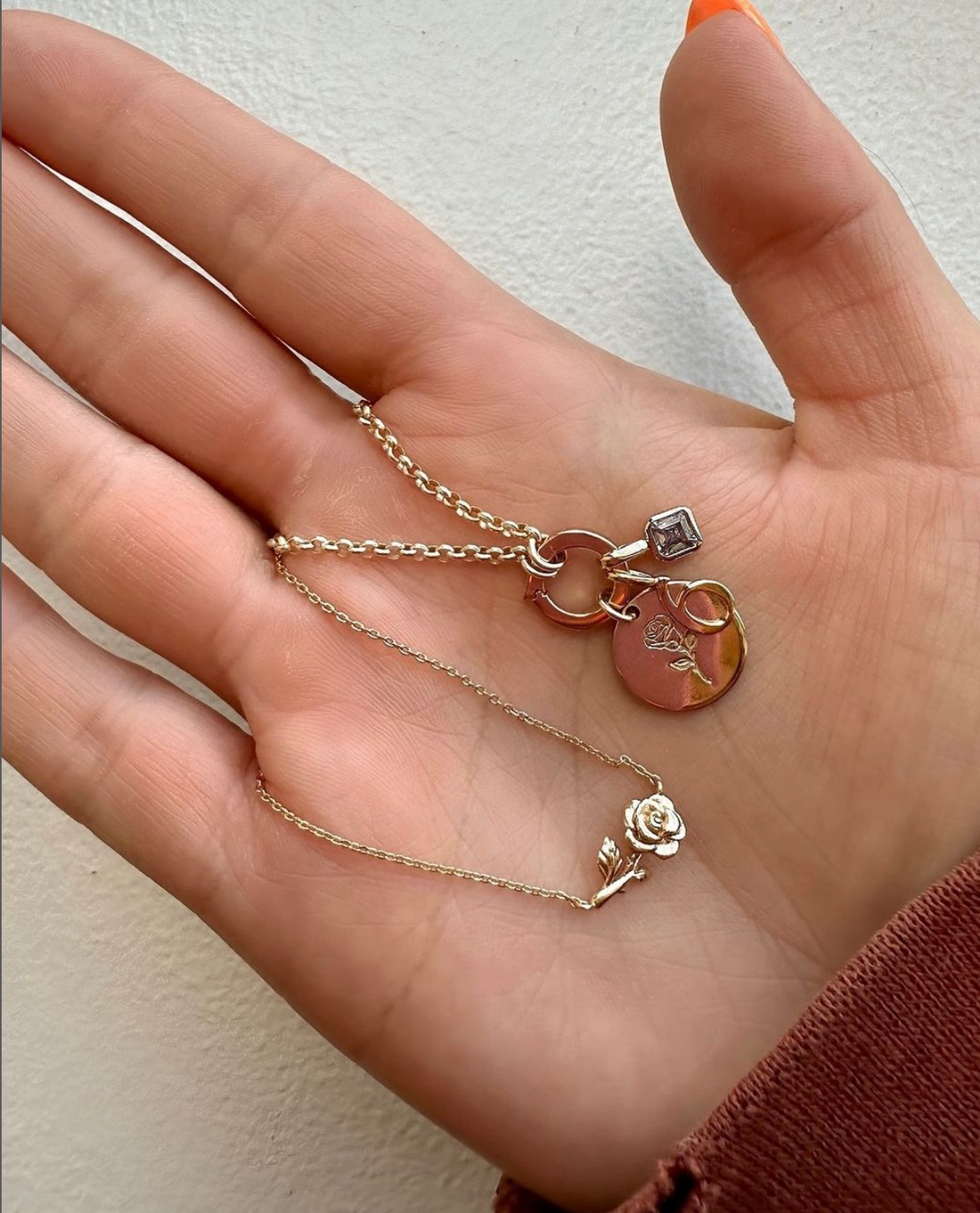 A gold charm necklace with a ring in the center, holding an engraved round disc charm, a number '6' charm, and a square gemstone charm.