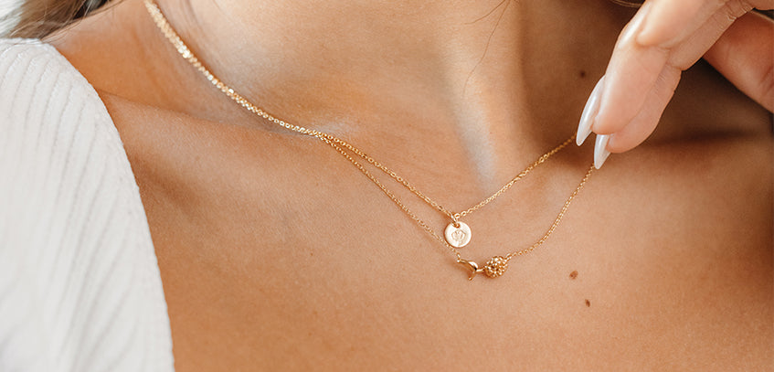 A woman wearing a delicate gold necklace with a horizontal flower stem in the center layered with a gold round disc pendant necklace.