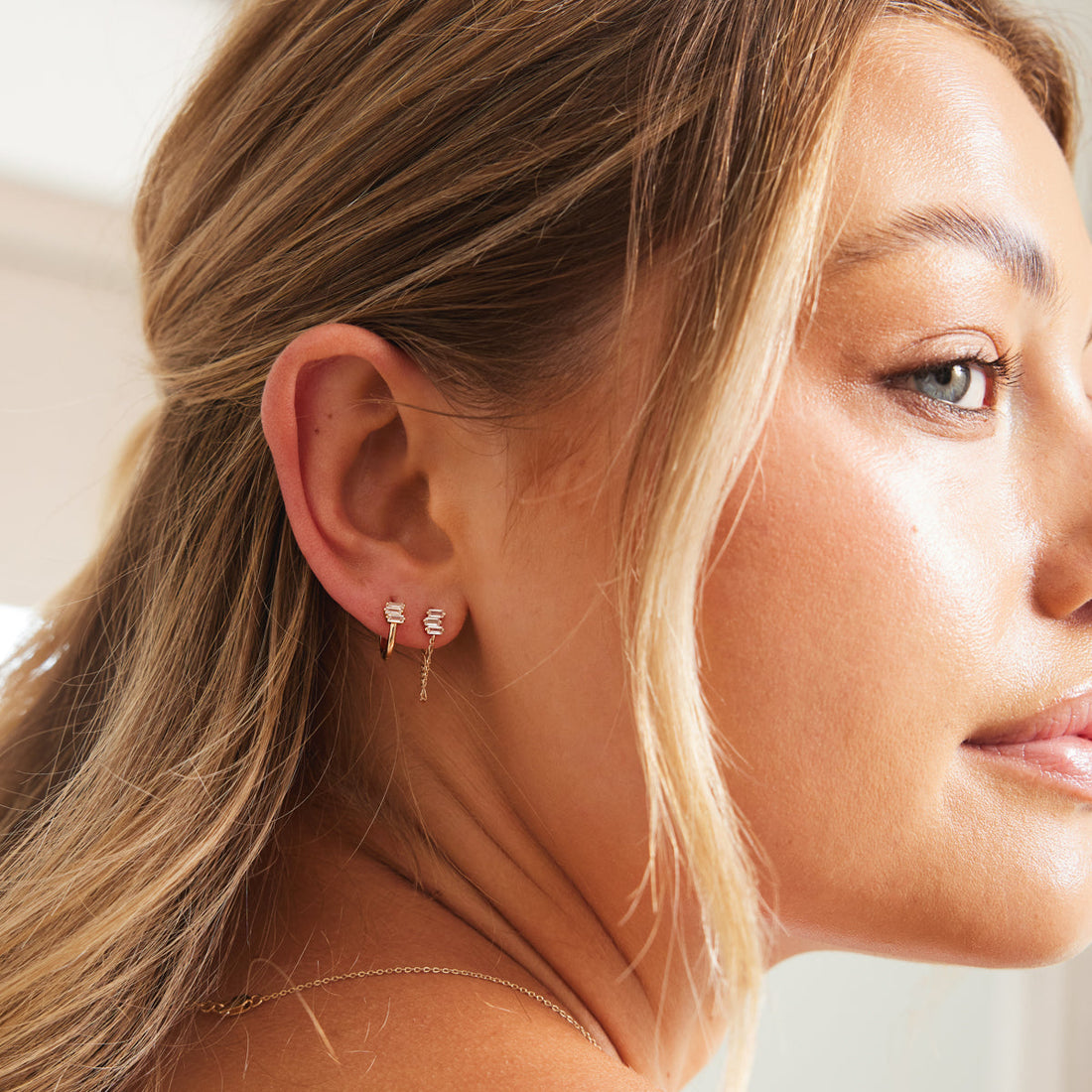 A woman wearing a gold hoop earring with three clear baguette stones, and the same style of baguette stone trio on a stud with a chain.