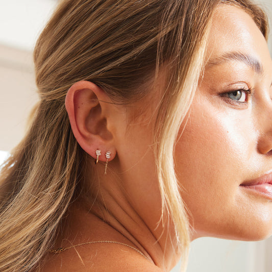 A woman wearing a gold hoop earring with three clear baguette stones, and the same style of baguette stone trio on a stud with a chain.