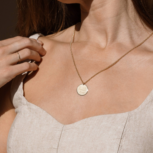 A person wearing a gold pendant chain necklace with a round disc, engraved with handwriting.