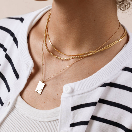 A woman wearing layered gold necklaces, including an engraved rectangle pendant necklace.
