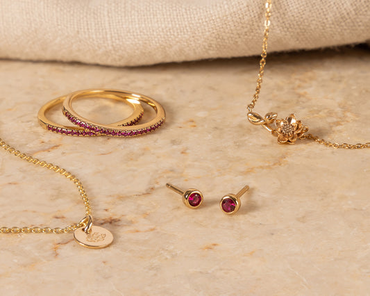 Multiple pieces of gold jewelry representing July with dark red gemstones and lotus flowers.