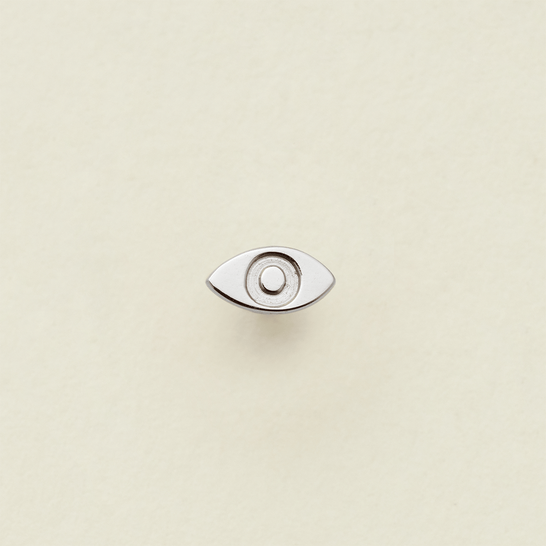 A silver stud earring in the shape of an eye, shown on a solid light beige surface.