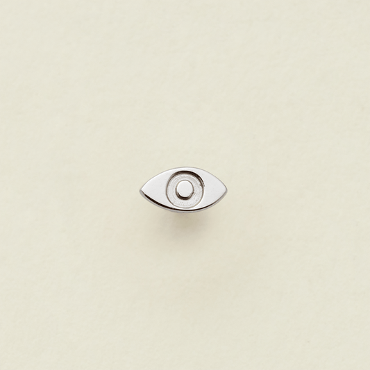 A silver stud earring in the shape of an eye, shown on a solid light beige surface.