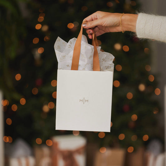 A cream colored Made by Mary gift bag with tissue paper, held in front of a Christmas Tree.