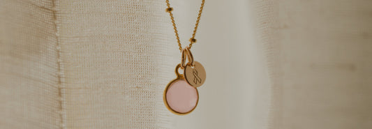 A gold pendant necklace with a pink gemstone charm and a small round disc charm engraved with the cancer ribbon symbol.