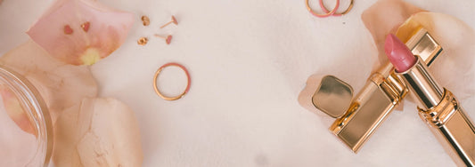 Gold and pink jewelry and a pink lipstick in a gold container laying on a marbled white surface.