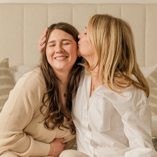 A woman kissing her adult daughter on the head, both wearing gold round disc pendant necklaces.