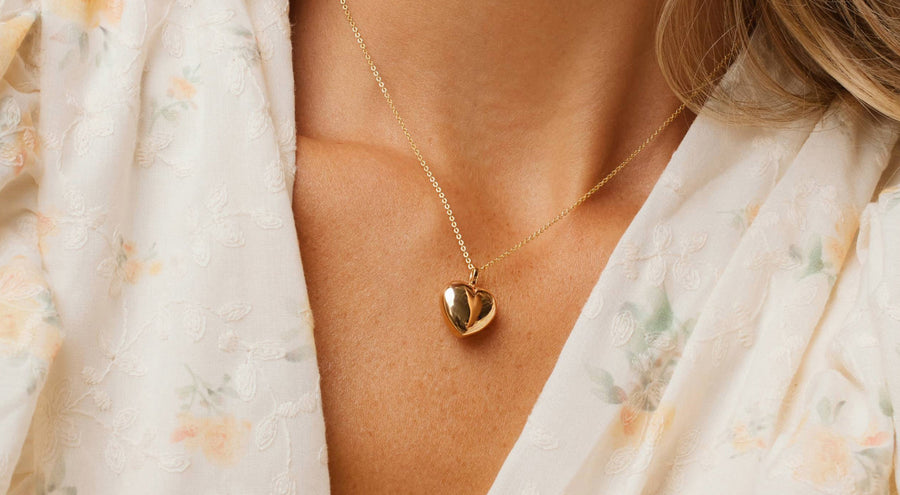 A person wearing a gold pendant chain necklace with a bold polished gold heart charm.