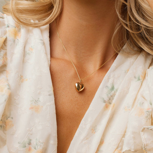 A person wearing a gold pendant chain necklace with a bold polished gold heart charm.
