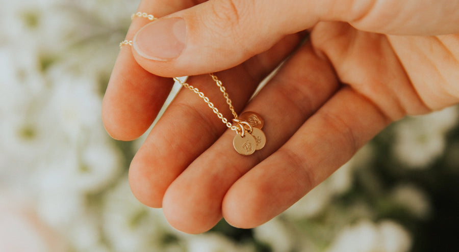 A person's hand in front of a bouquet of white flowers holding a gold pendant chain necklace with multiple small engraved disc charms.
