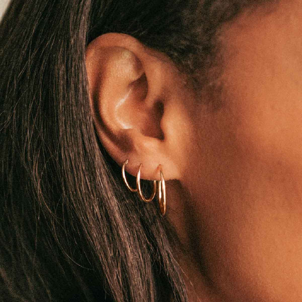 A close-up of a person's ear wearing multiple sizes of thin gold wire hoop earrings.