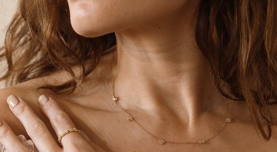 A person wearing a gold necklace and ring, both adorned with small stationary floral charms.