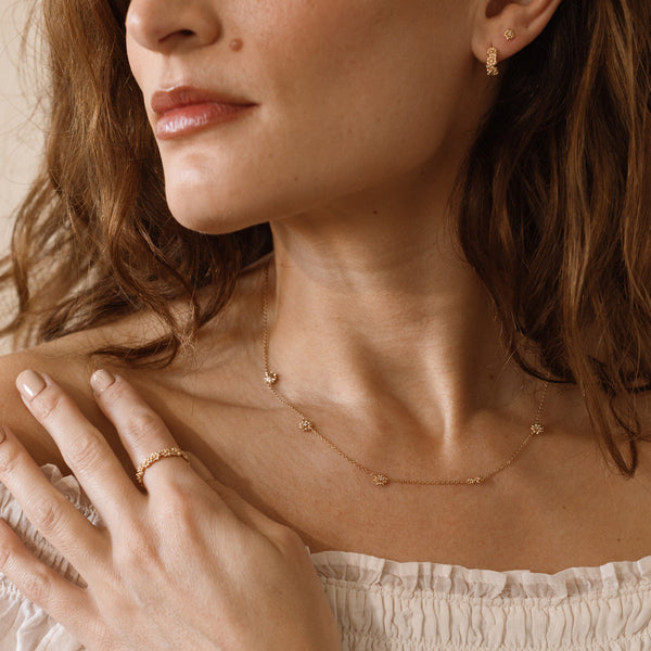 A person wearing a gold necklace and ring, both adorned with small stationary floral charms.