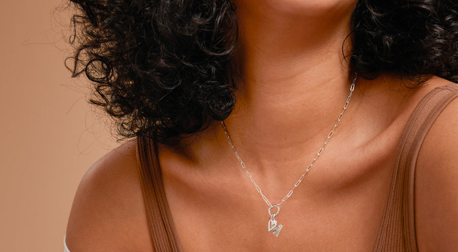 A close-up of a woman in a light brown tank top wearing a silver paperclip chain charm necklace with multiple silver charms.