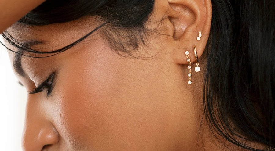 A person's ear wearing multiple gold earrings in different styles, each embellished with clear round gemstones.
