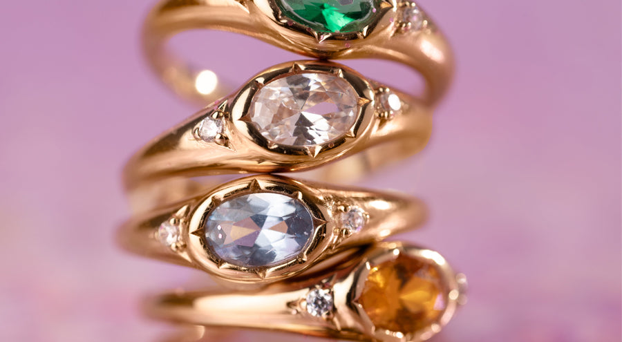 Four stacked gold rings on a pink background, each with a bezel set oval gemstone in a different color.