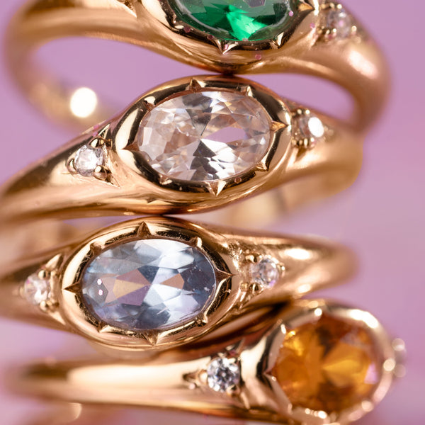 Four stacked gold rings on a pink background, each with a bezel set oval gemstone in a different color.