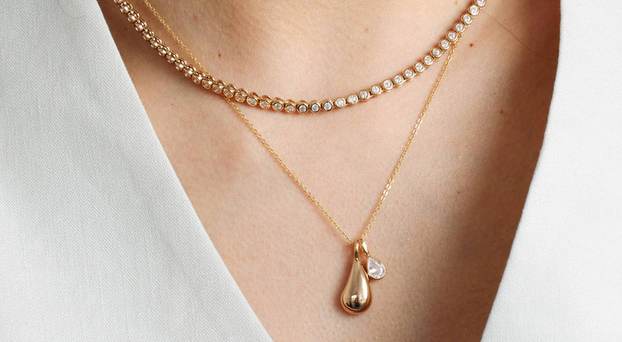 A person wearing a gold tennis necklace layered with a long gold pendant chain with a polished gold tear drop charm.
