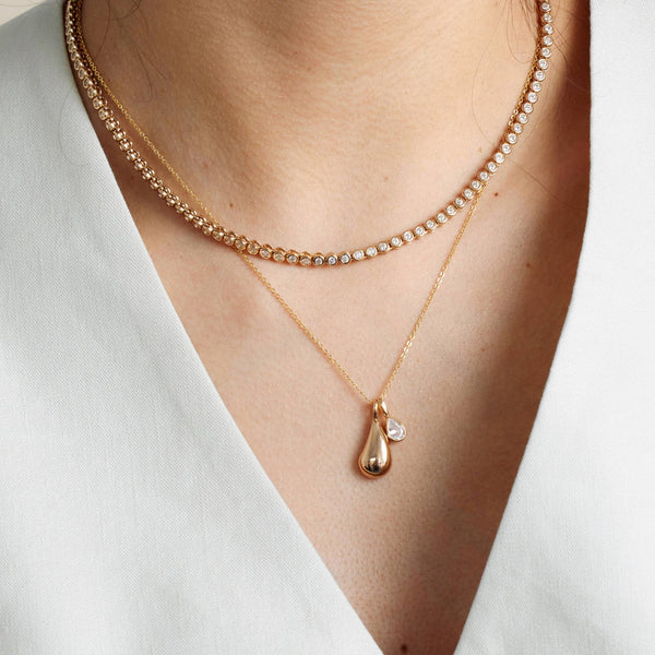 A person wearing a gold tennis necklace layered with a long gold pendant chain with a polished gold tear drop charm.