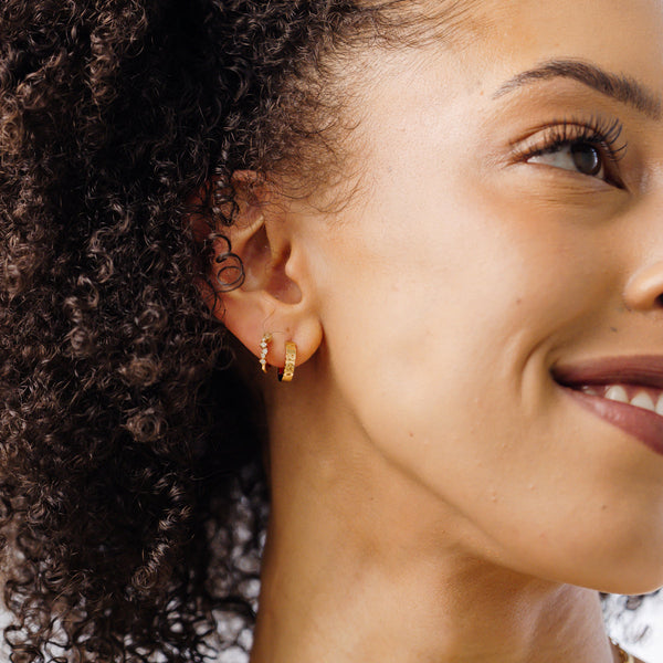 A close-up of a woman's ear wearing two pairs of gold huggie hoops, one covered with small clear accent stones.