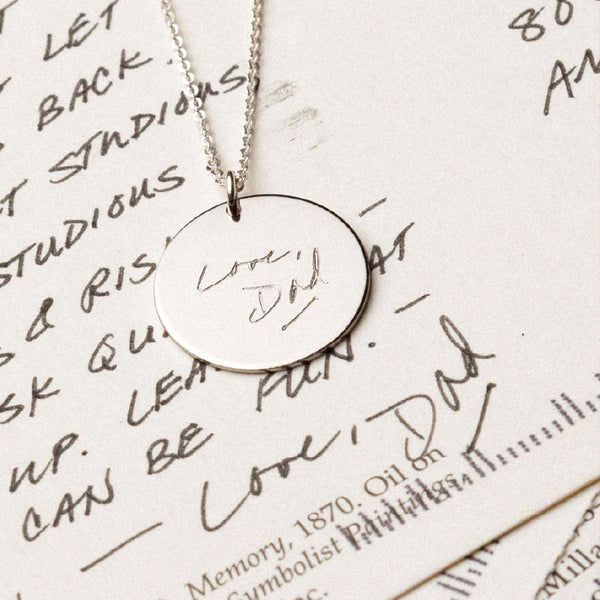 A silver round disc pendant necklace engraved with text, laying on a black and white handwritten letter.