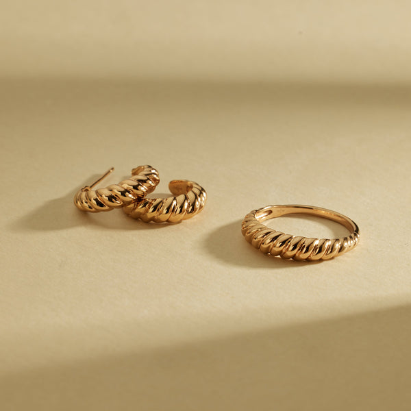 A pair of textured gold hoop earrings and a matching bold gold ring, laying on a light beige surface.