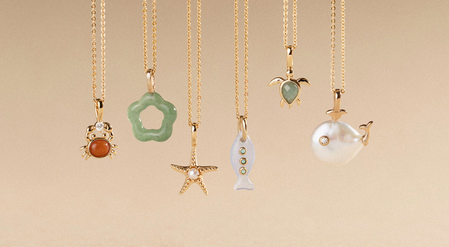 Six gold pendant necklaces, each with a different ocean inspired charm, hanging in front of an ombre beige background.