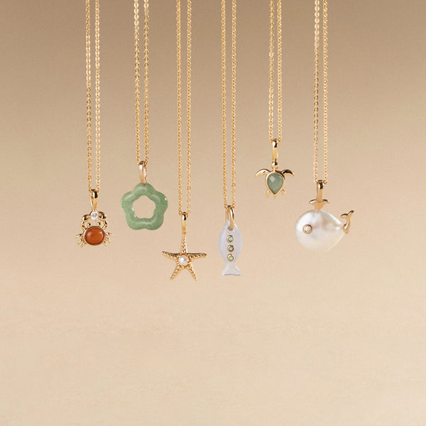 Six gold pendant necklaces, each with a different ocean inspired charm, hanging in front of an ombre beige background.