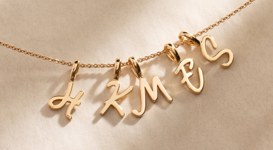 A gold pendant necklace with an initial H, K, M, E, and S charm in script font.