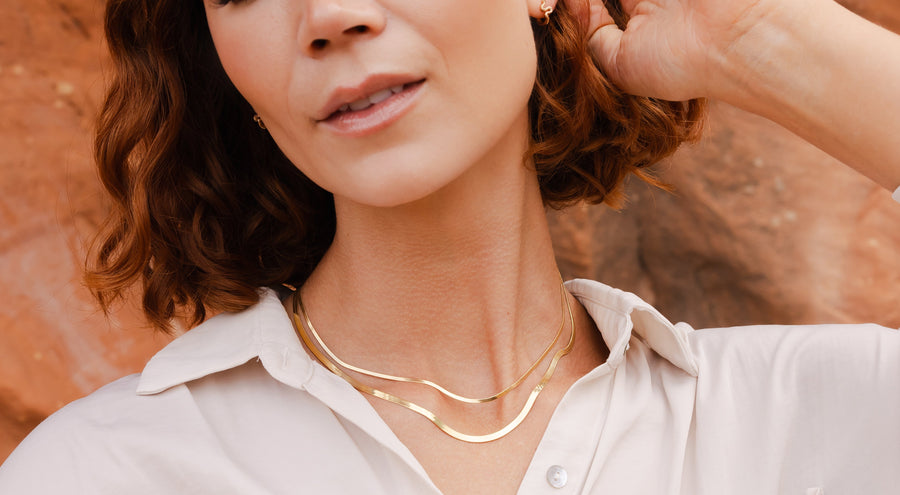 A model wearing a white button-up top standing in front of red rocks, wearing multiple pieces of gold jewelry.