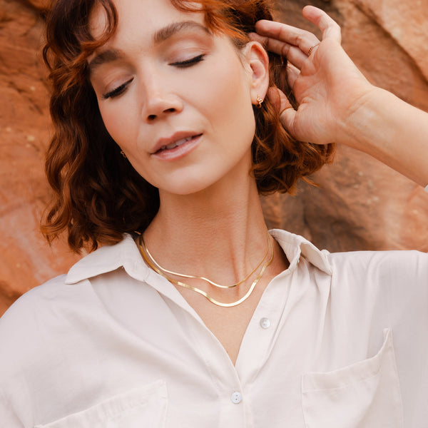 A model wearing a white button-up top standing in front of red rocks, wearing multiple pieces of gold jewelry.