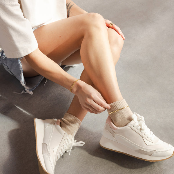 A person's legs wearing white tennis shoes and light brown socks, paired with a gold chain anklet.