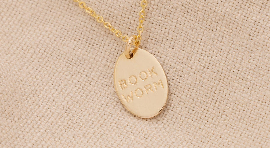 A delicate gold pendant chain necklace with a small oval charm engraved with the phrase 'BOOK WORM'.