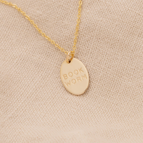 A delicate gold pendant chain necklace with a small oval charm engraved with the phrase 'BOOK WORM'.